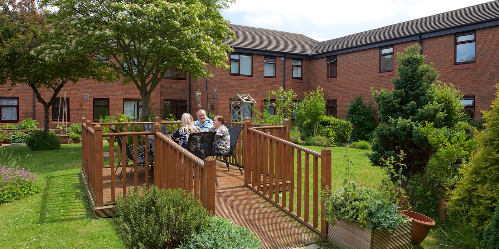 A group of people sitting outside on a wooden deck surrounded by green gardens, with a home in the background