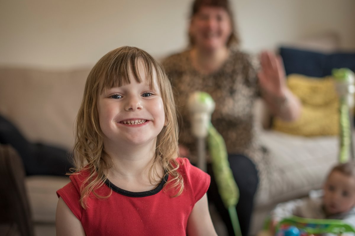 A young child in a red teeshirt smiling broadly from the inside of her home