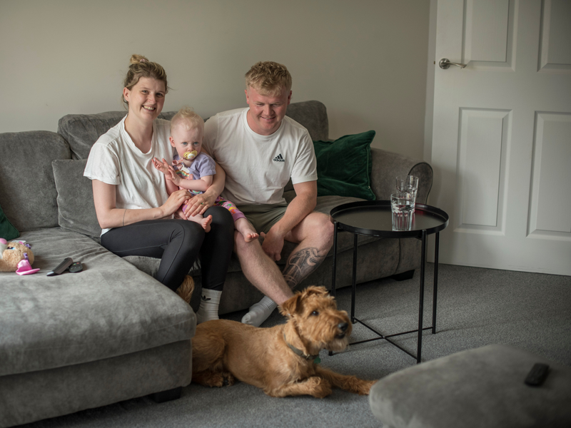 A family of three sitting on a grey sofa with a shaggy brown dog at their feet